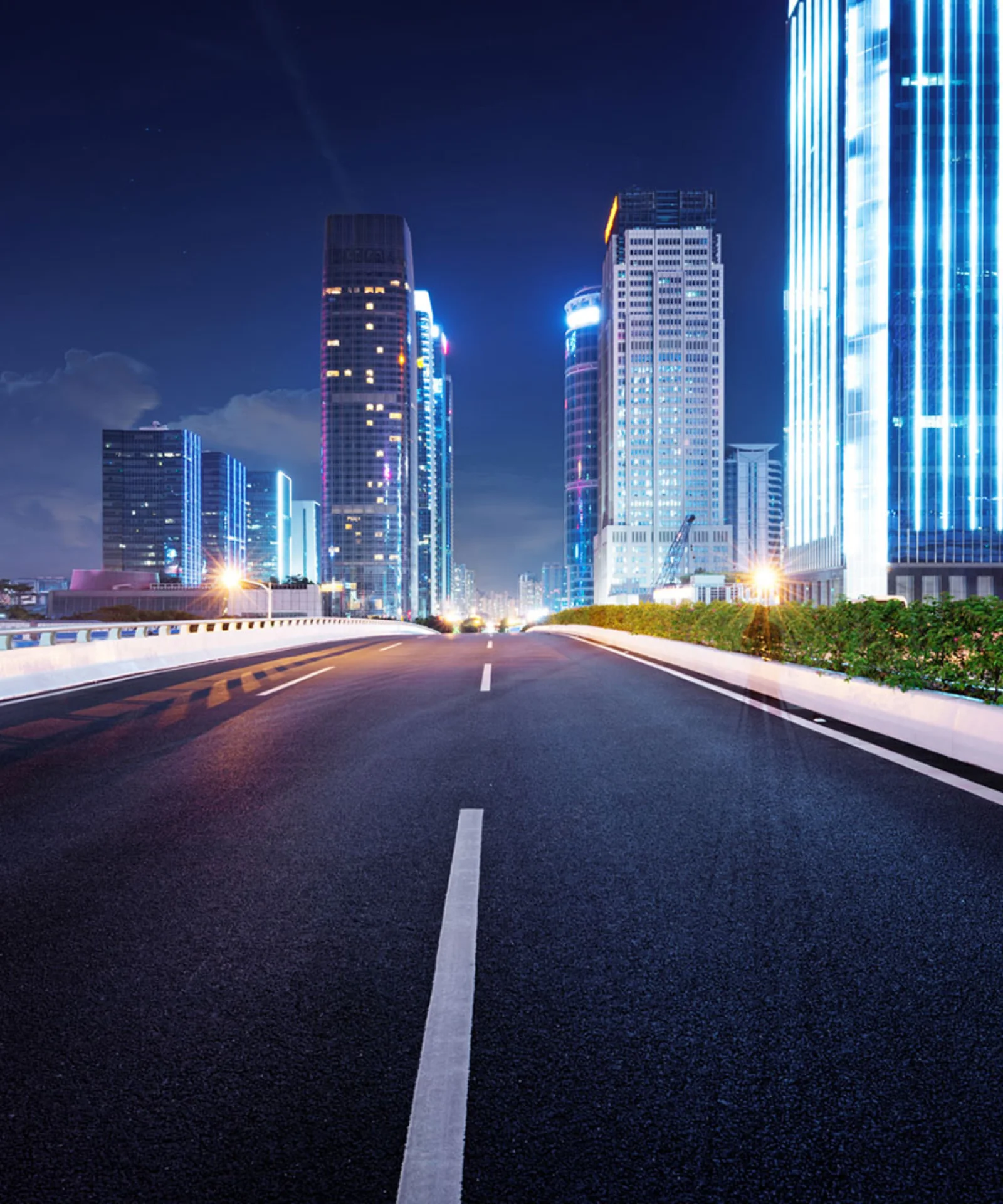 Illuminated cityscape at night with an empty highway leading towards skyscrapers.