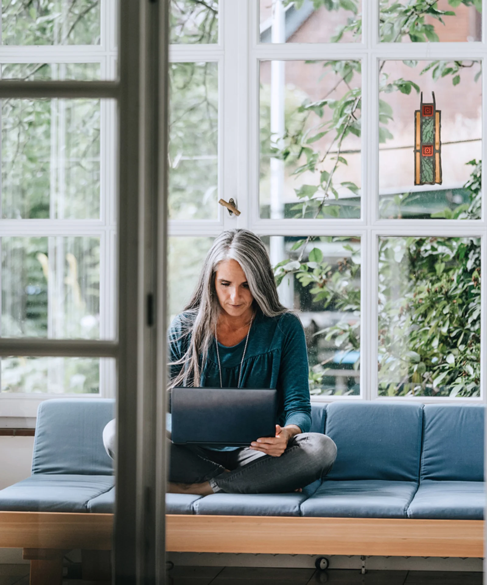 Woman working on a laptop while sitting on a couch in a bright room.