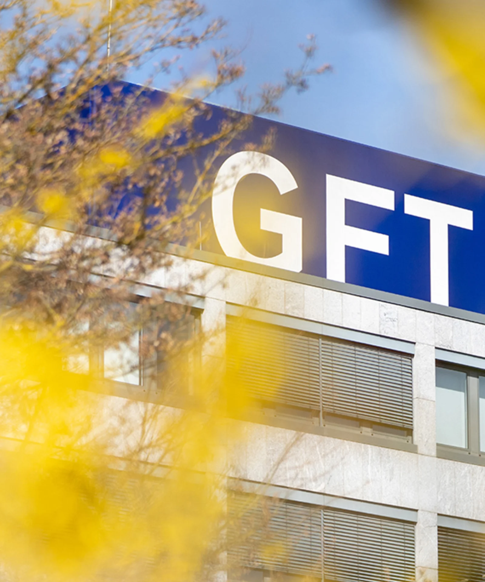 Edificio de la sede de GFT con el logotipo de la empresa destacado en la parte superior, enmarcado por un follaje amarillo vibrante en primer plano.