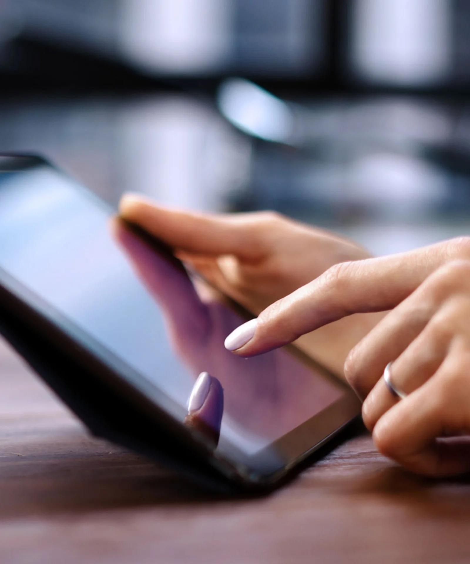 The image shows a close-up of hands using a tablet, with one hand tapping on the screen. The tablet is lying on a surface and the background is slightly blurred, suggesting an interior space. The focus is on the interaction with the tablet.