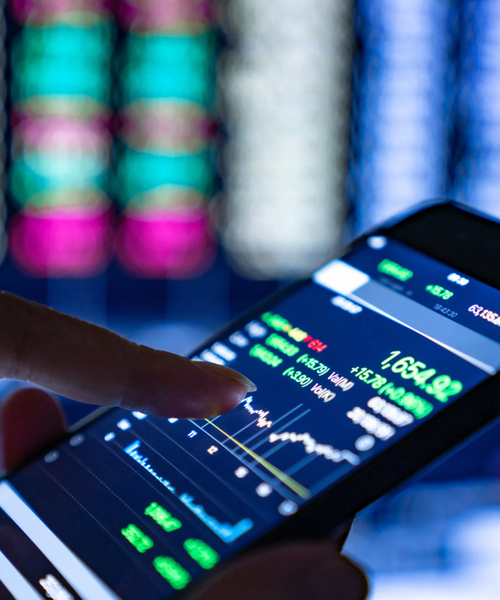 A close-up of a hand using a smartphone to monitor stock market data with blurred financial screens in the background.