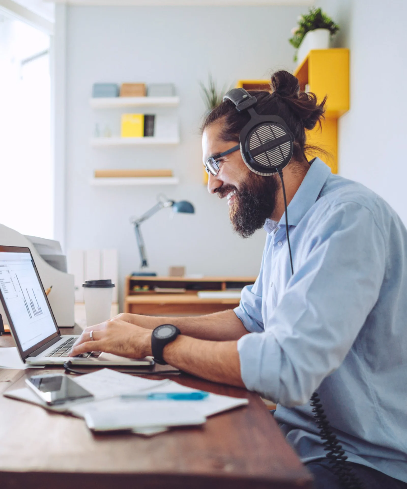 Man wearing headphones and glasses working on a laptop at home office, surrounded by tech gadgets and documents.