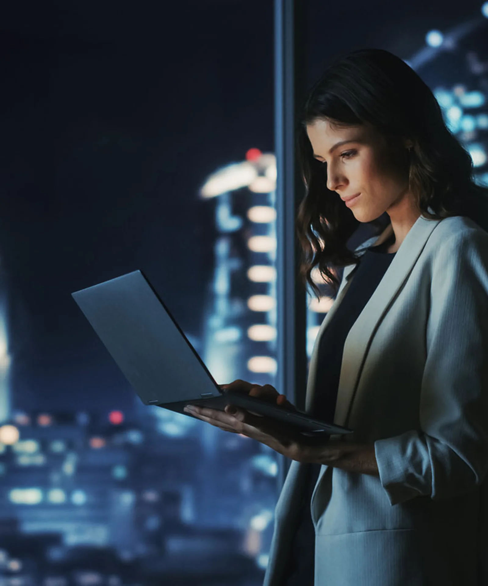 Female professional working on a laptop in a high-rise office at night, with a city skyline illuminated in the background.