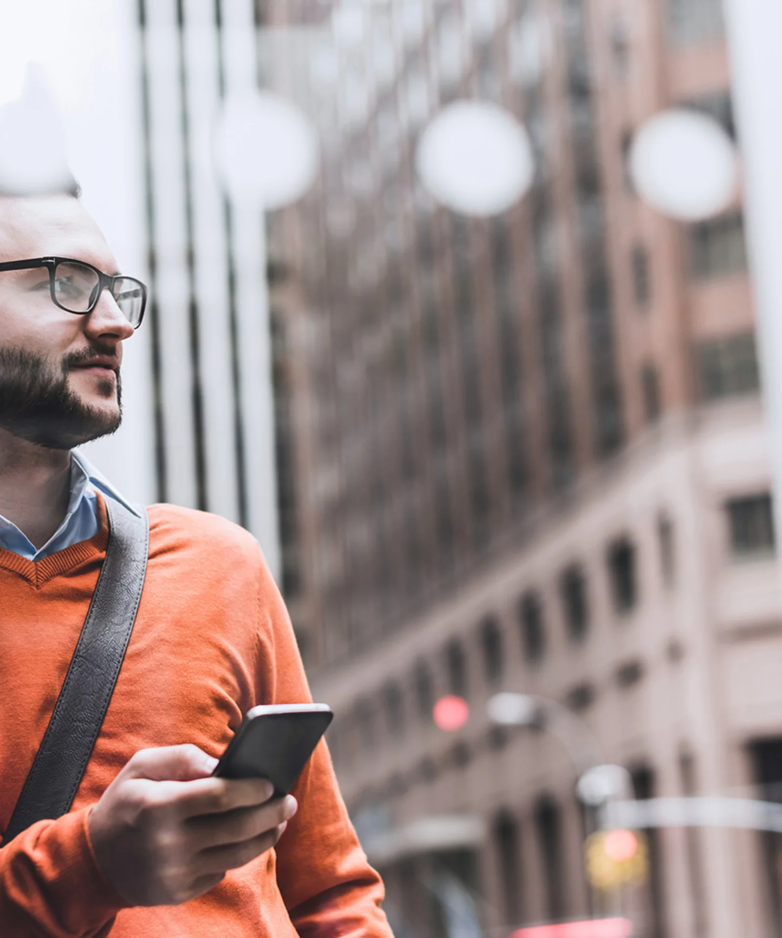 Mann im orangefarbenen Pullover hält ein Smartphone und steht in einer städtischen Umgebung.