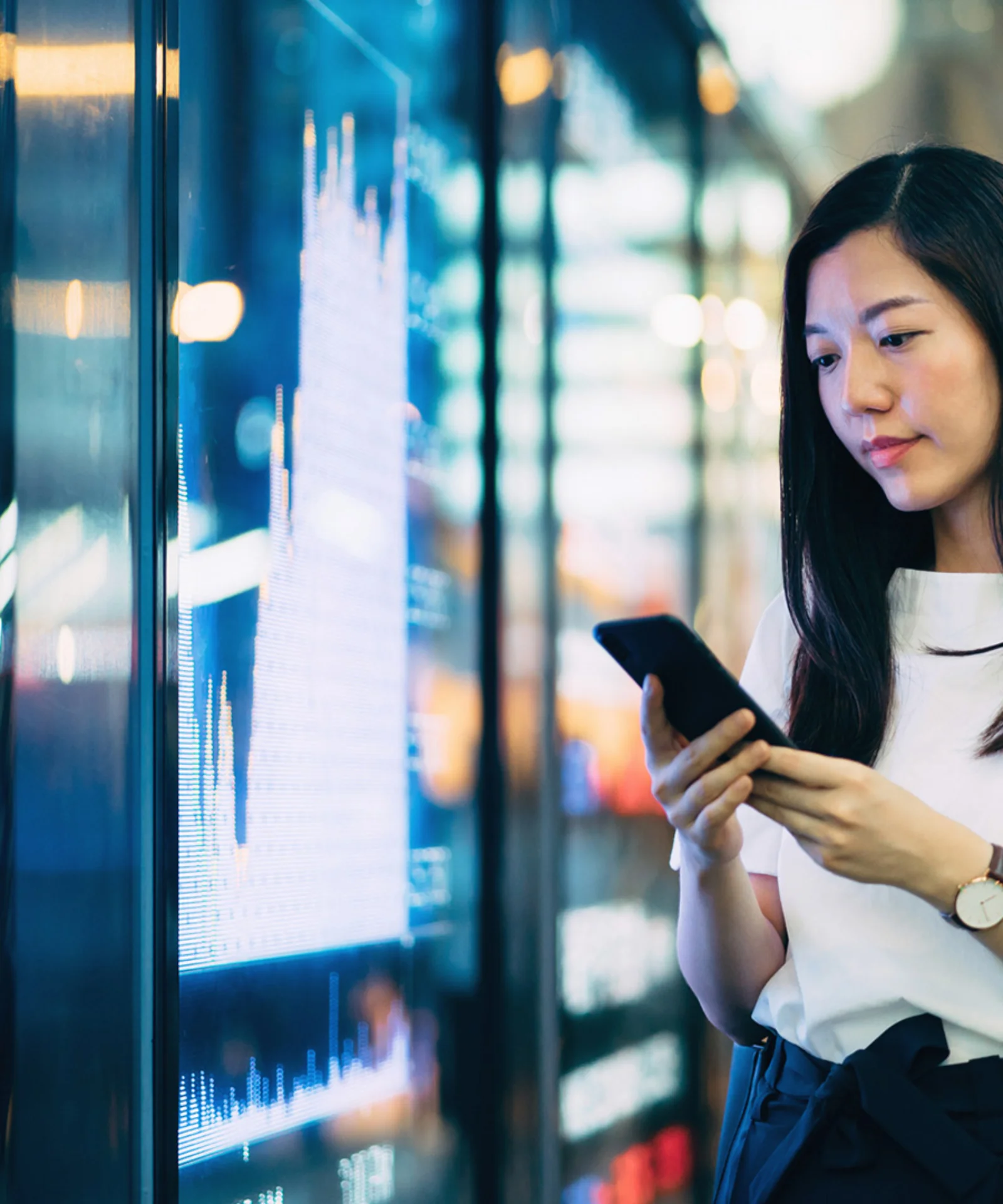 imagem mostra uma mulher usando seu smartphone enquanto está em pé na frente de um quadro eletrônico que exibe dados financeiros. Este visual simboliza a integração da sustentabilidade em tecnologia e práticas comerciais, destacando a importância do desenvolvimento sustentável na era digital moderna. O uso da tecnologia no monitoramento e promoção de iniciativas sustentáveis é um tema-chave representado nesta imagem.