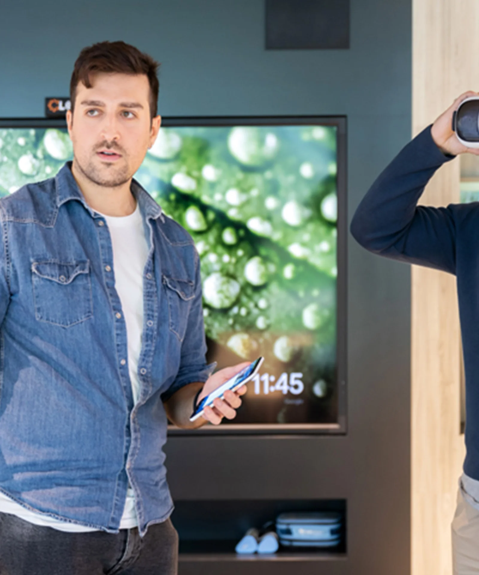 This image captures an interactive session in an innovation lab where two men are engaging with advanced technologies. One man is wearing a virtual reality headset, experiencing immersive content, while the other man is holding a smartphone and possibly explaining or presenting something. The background features a large screen displaying vivid visuals, enhancing the modern and tech-centric ambiance of the innovation lab. This setting reflects the integration of cutting-edge technologies and collaborative environments aimed at fostering innovation and creativity.
