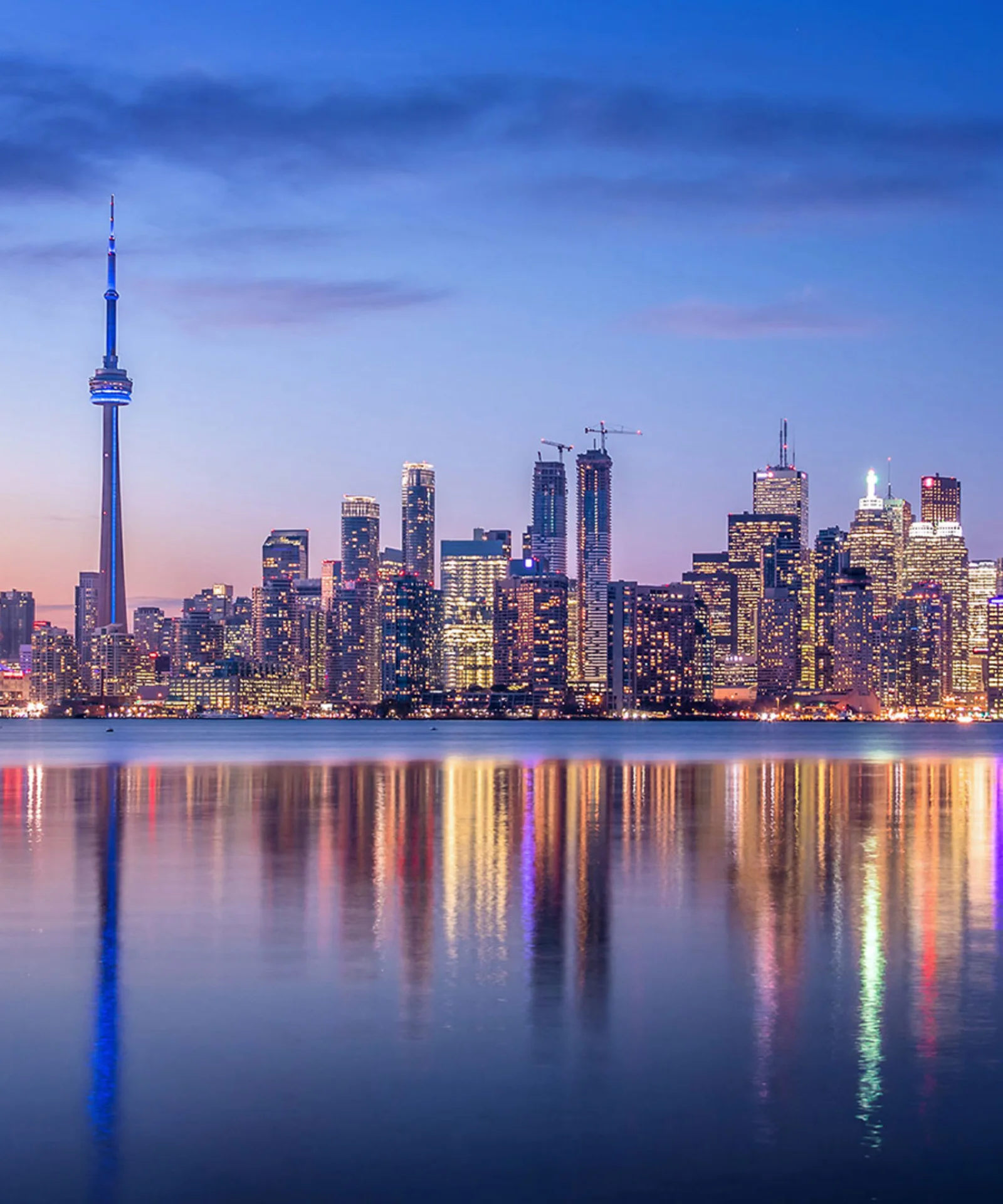 The image features a stunning city skyline at dusk, with tall buildings and the iconic CN Tower prominently visible. The serene water in the foreground reflects the vibrant lights of the city, creating a symmetrical and tranquil scene. This visual captures the essence of stability, growth, and the future, making it an ideal representation for the insurance industry. The clear, calm waters and the well-lit cityscape symbolize the industry&#039;s promise of security, protection, and forward-thinking solutions.