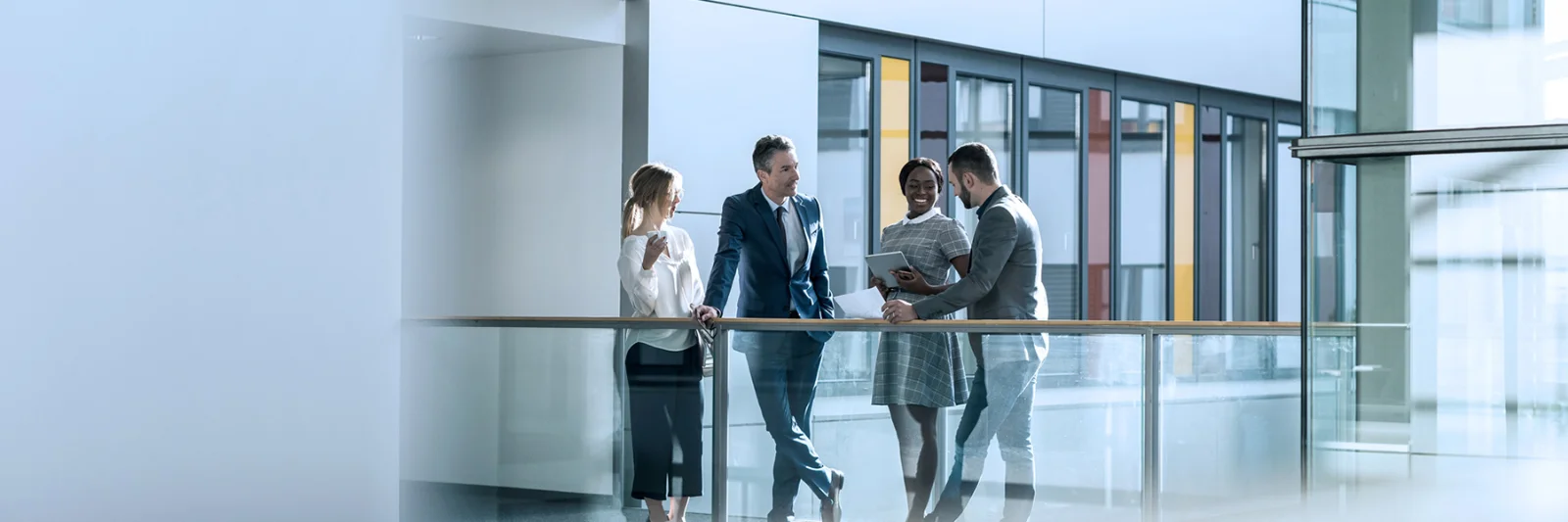 This image captures a professional meeting in a modern office setting. Four individuals, three men and one woman, are engaged in a conversation, standing by a glass railing on an upper floor. The office environment features clean, contemporary design elements with large windows and glass partitions, creating a bright and open atmosphere. The people are dressed in business attire, suggesting a formal and collaborative business environment. This visual is ideal for conveying partnership, collaboration, and a modern business ethos.