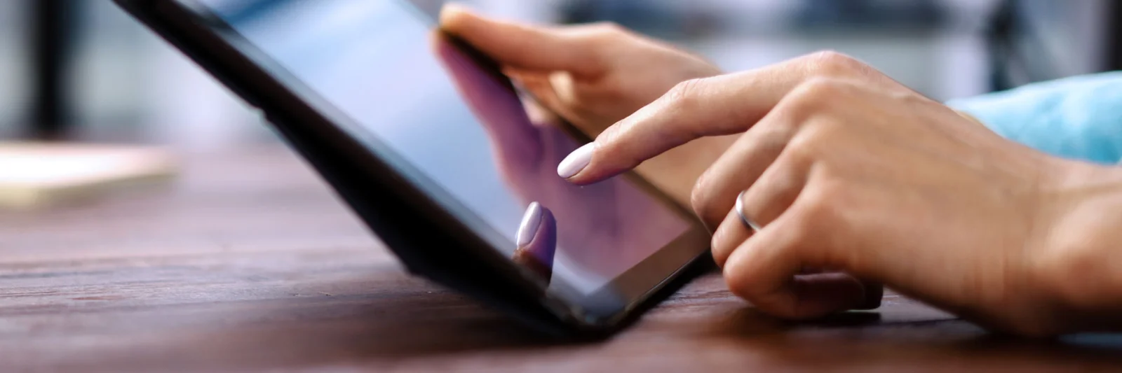 The image shows a close-up of hands using a tablet, with one hand tapping on the screen. The tablet is lying on a surface and the background is slightly blurred, suggesting an interior space. The focus is on the interaction with the tablet.