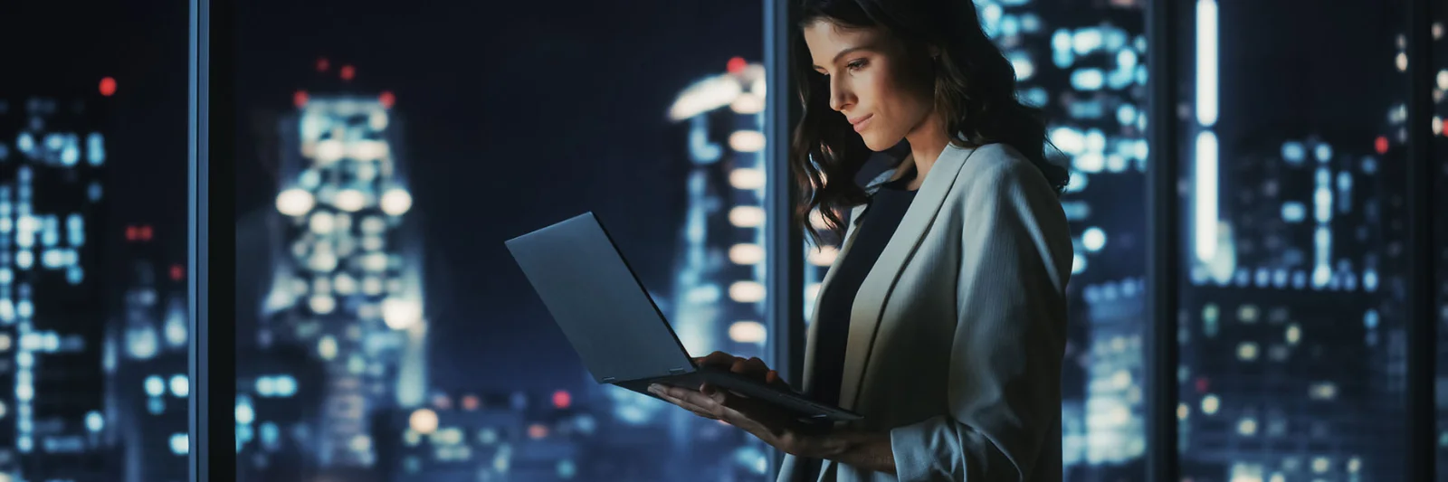 Female professional working on a laptop in a high-rise office at night, with a city skyline illuminated in the background.