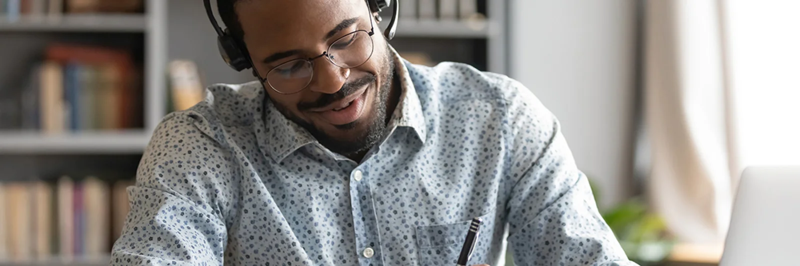 A man with headphones taking notes while listening to a podcast on his laptop, representing GFT&#039;s podcast library. He is seated in a cozy home office with bookshelves in the background, showcasing a comfortable and productive environment for learning and engagement.