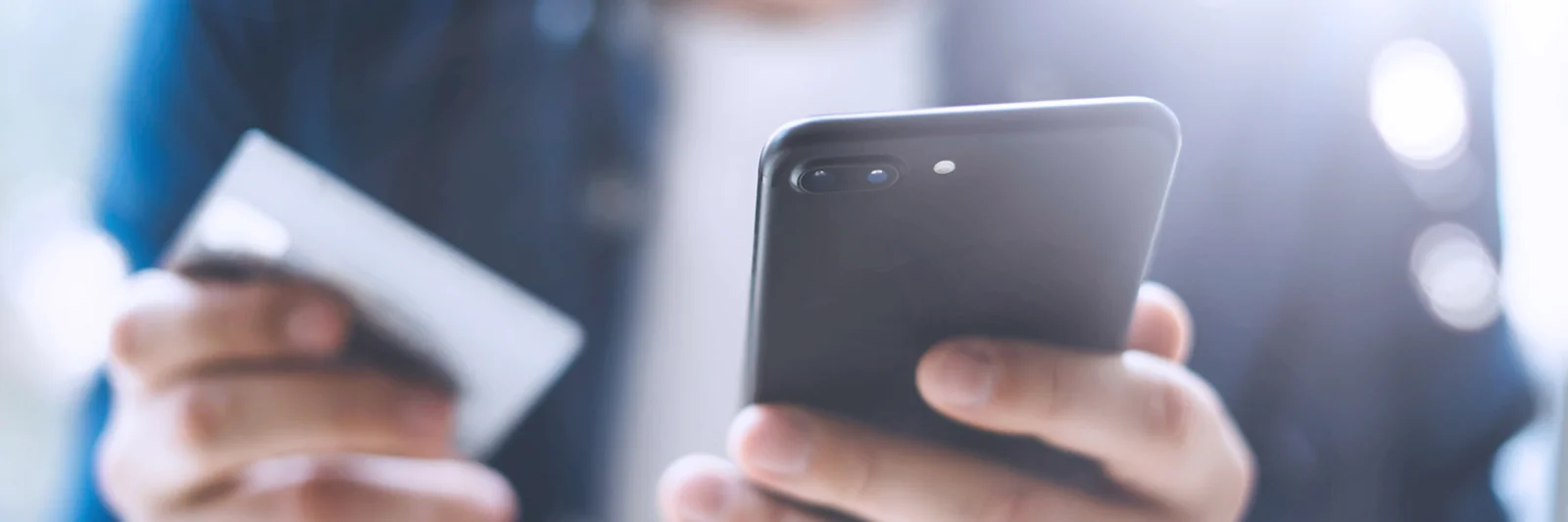 Close-up of a man holding a smartphone in one hand and a credit card in the other, representing mobile payment and online shopping.