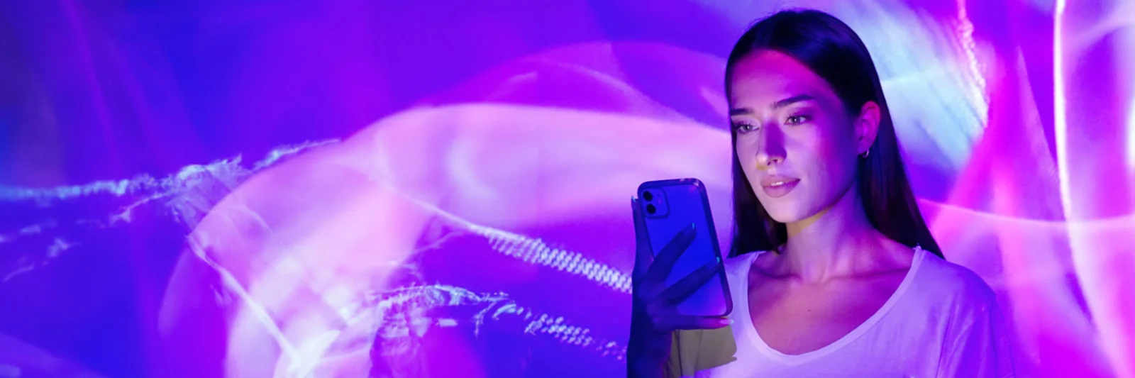This key visual for GFT Banking Agent showcases a woman holding a smartphone, enveloped in a mesmerizing display of abstract purple and pink lights. The image evokes a sense of modern technology and digital innovation, reflecting the dynamic and futuristic nature of the GFT Banking Agent. The interplay of lights and shadows creates an immersive and visually captivating atmosphere, symbolizing the seamless integration of advanced AI and banking technology in enhancing customer experiences.