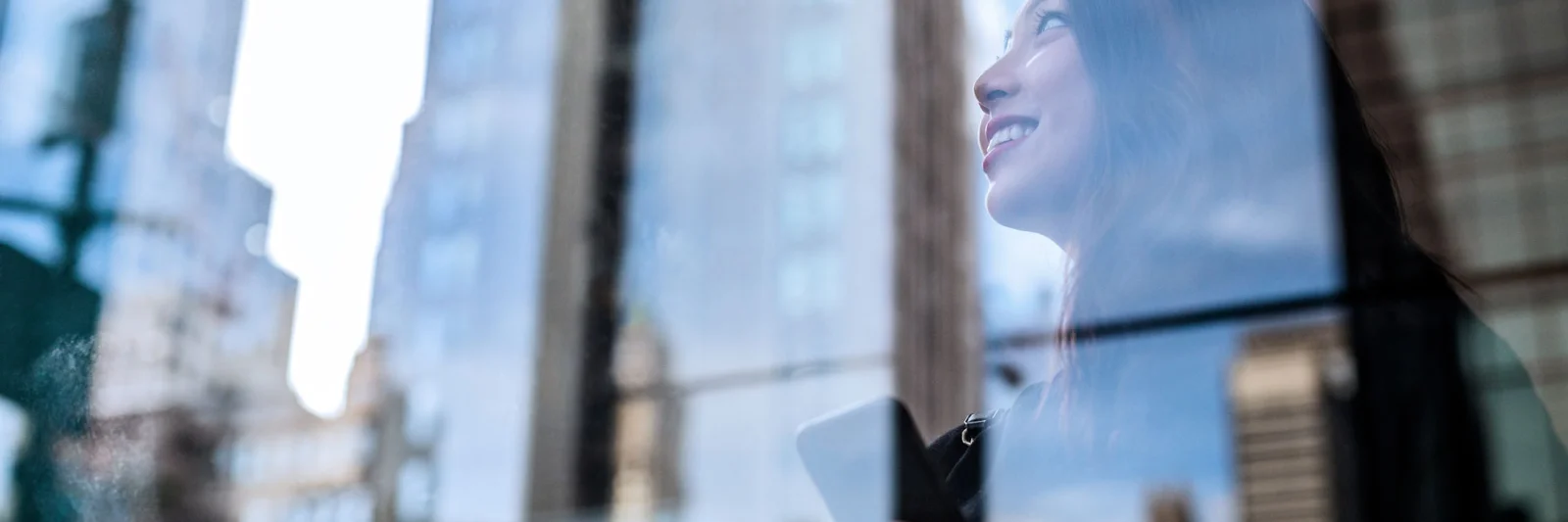 Una mujer sosteniendo un teléfono inteligente, sonriendo mientras mira hacia arriba, con el reflejo de los rascacielos en una ventana de vidrio, simbolizando la innovación y el crecimiento.