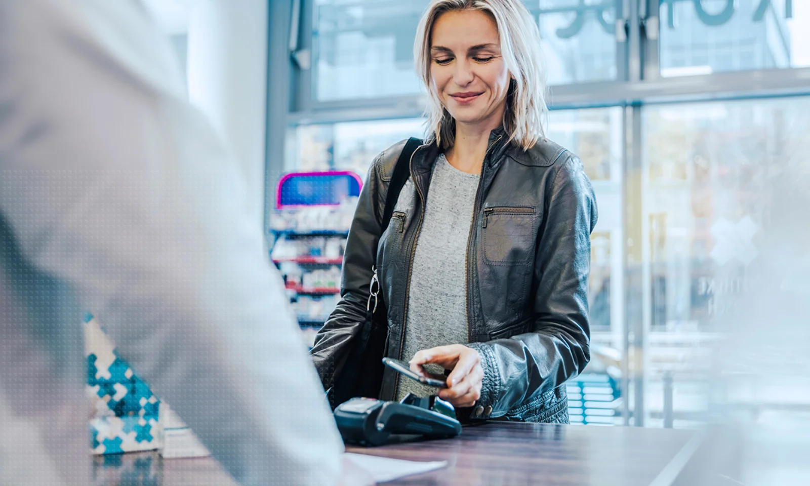 A woman in a leather jacket makes a contactless payment with her smartphone at a retail store counter, demonstrating modern digital payment technology.