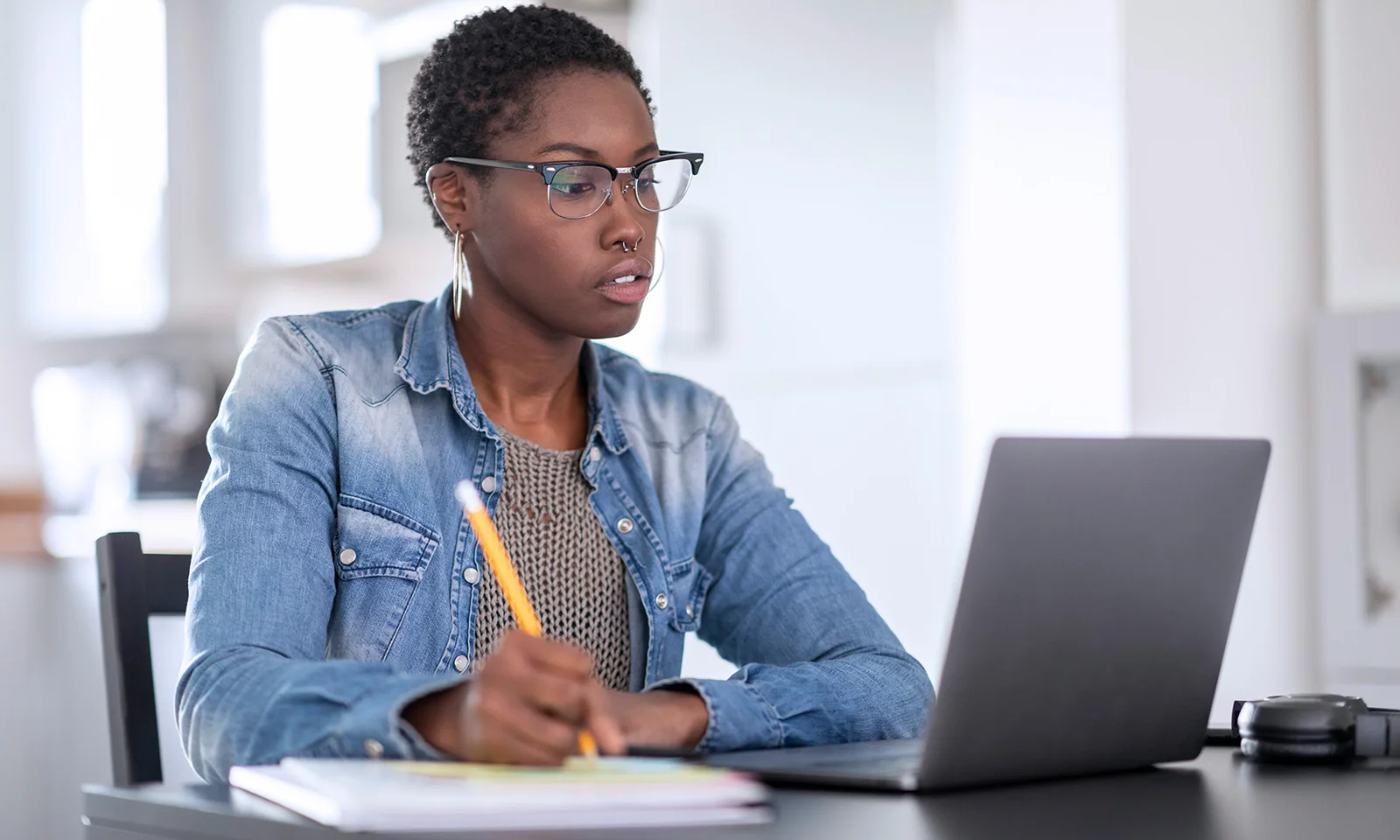 Focused professional woman working remotely on a laptop, taking notes with a pencil in a bright home office.