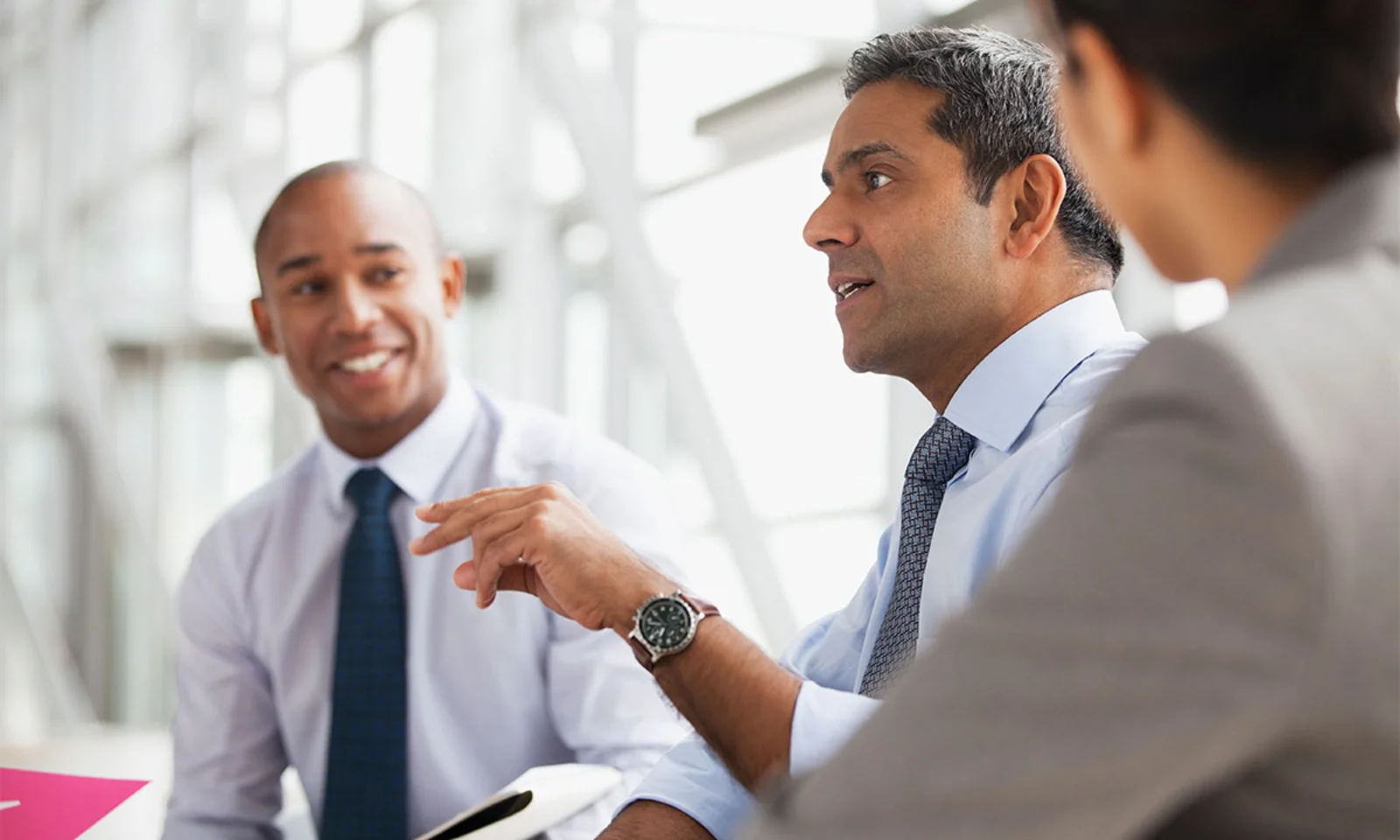 Three business professionals engaged in a collaborative discussion in a modern office setting, with natural light streaming through large windows.