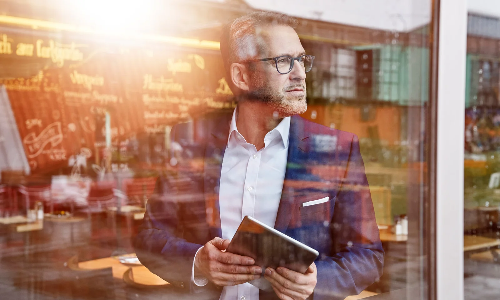Business professional holding a tablet, gazing thoughtfully through a cafe window. This image captures GFT&#039;s dedication to digital transformation and strategic innovation, showcasing how technology drives business success in modern environments.