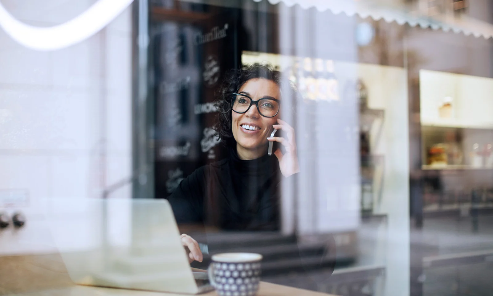 A professional woman smiling while working on her laptop and talking on the phone in a café, representing the flexibility of modern work environments.