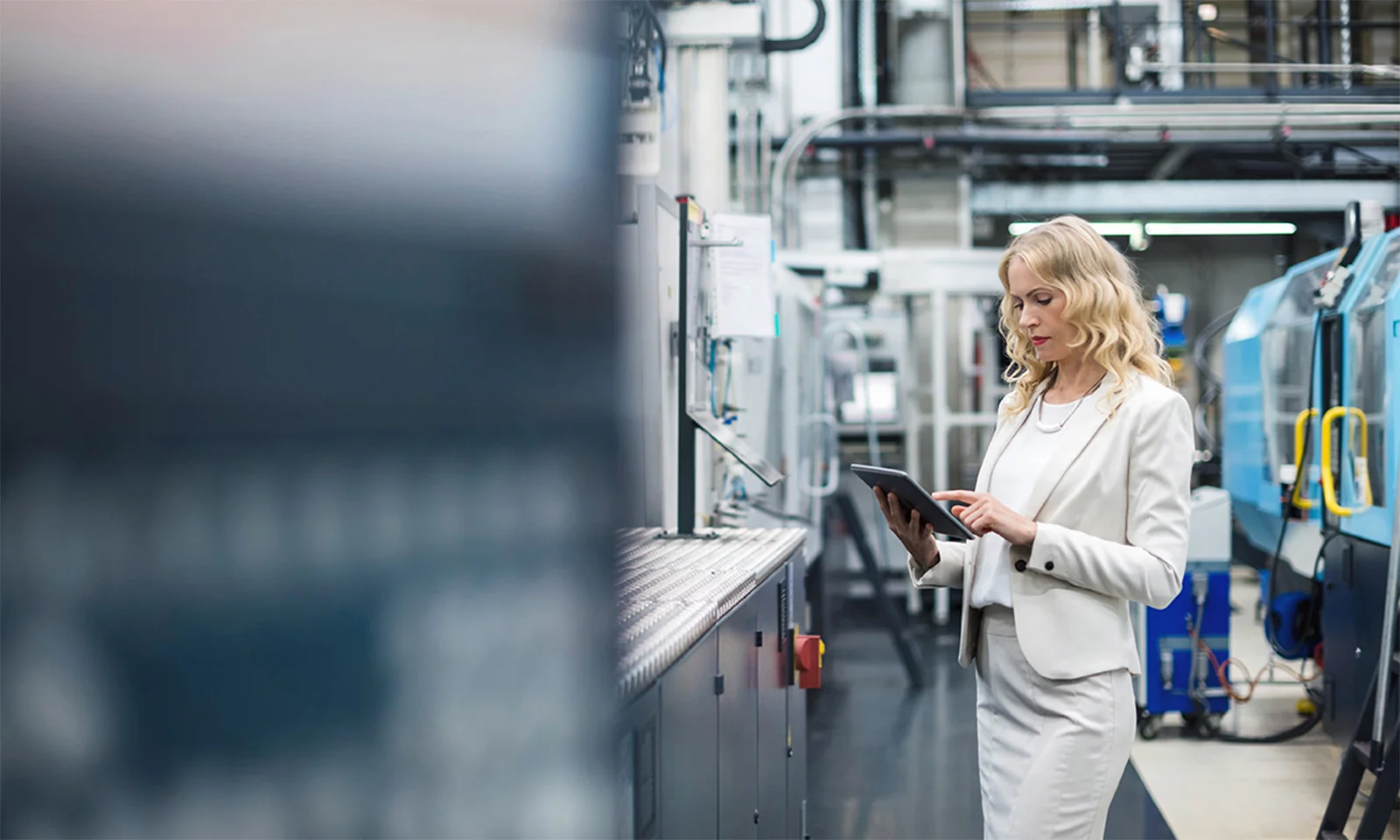 This image portrays a professional woman in a smart factory environment, using a tablet to monitor and manage production processes. She is dressed in a light-coloured suit, standing amidst advanced machinery and equipment, highlighting the integration of modern technology in sustainable manufacturing. The setting illustrates the commitment to sustainability in production, showcasing how smart factories utilise digital tools to enhance efficiency, reduce waste, and optimise resource usage. This visual emphasises the role of digitalisation in achieving sustainable production goals.