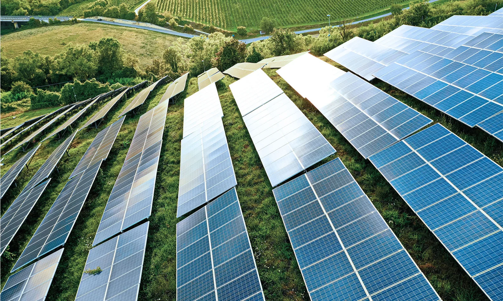 This image showcases a vast array of solar panels installed on a lush, green hillside, representing the concept of smart energy management and energy efficiency. The solar panels are strategically aligned to maximise sunlight capture, symbolising the use of renewable energy sources. The background features verdant fields and a winding road, further emphasising the harmony between advanced technology and the natural environment. This visual illustrates the integration of sustainable energy solutions in managing and reducing energy consumption, promoting a cleaner and more efficient energy future.
