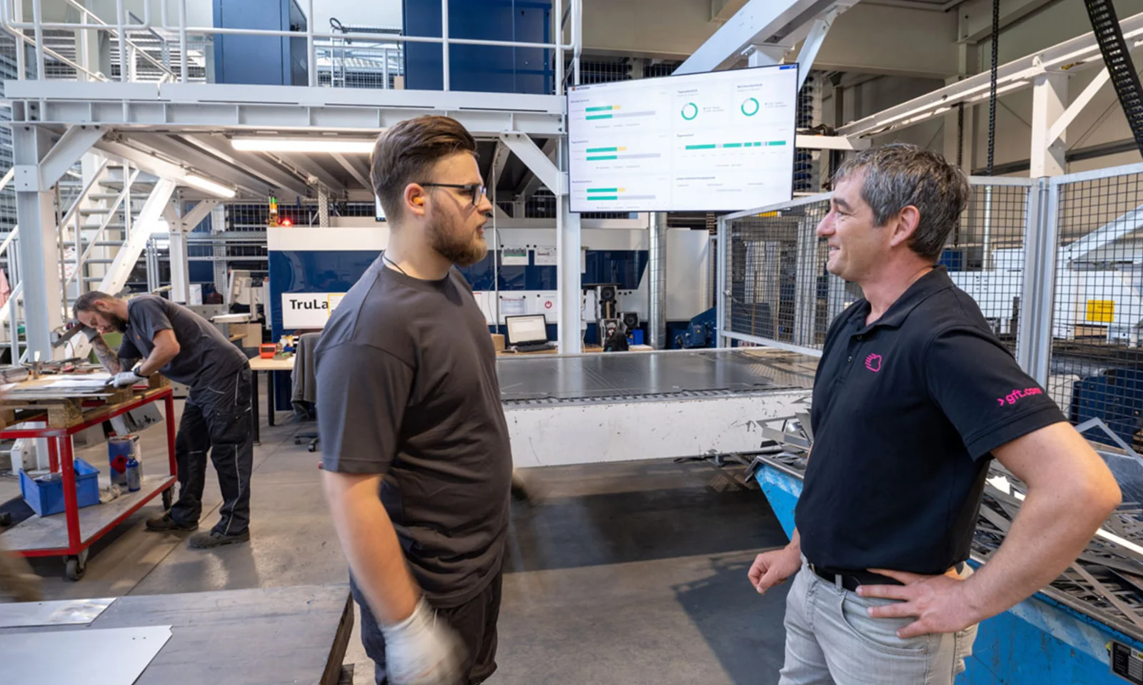 Two employees engage in a discussion on the shopfloor, with real-time data displayed on a large screen in the background, illustrating transparent and data-driven management at Schinko.