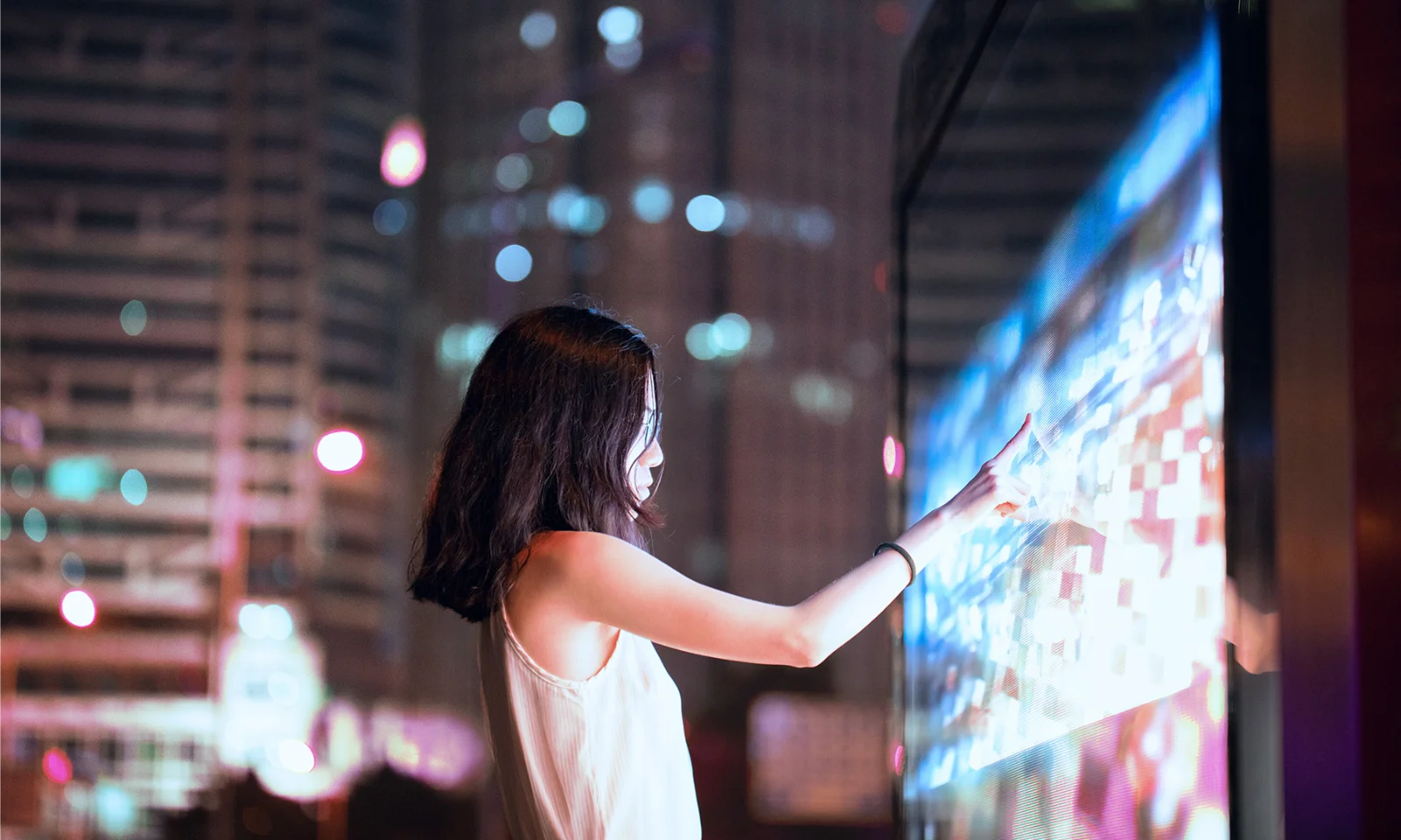 Young woman interacting with a large digital touch screen in a vibrant city at night, surrounded by bright lights and modern architecture.