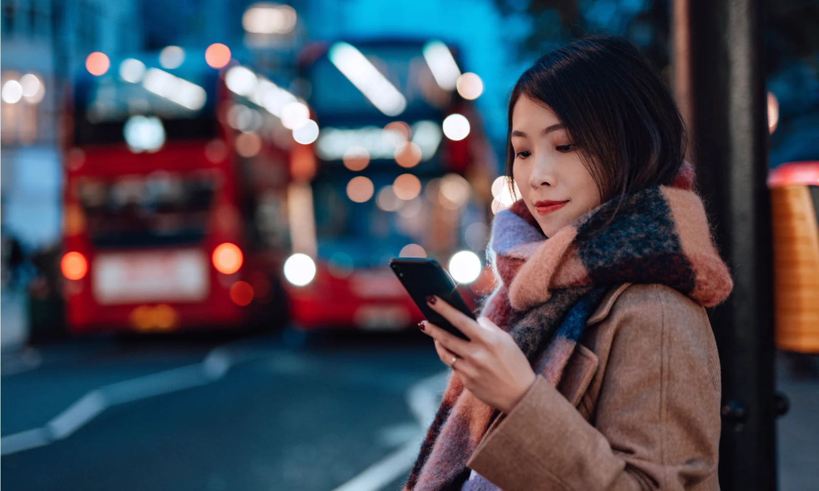 A woman in a city setting using her mobile phone, with blurred double-decker buses in the background, symbolising the integration of digital services in everyday urban life.