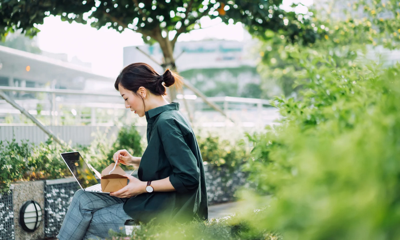 The image depicts a woman working on a laptop outdoors, surrounded by lush greenery, while eating from a takeaway box. This scene symbolizes the integration of green practices into daily work life, highlighting the balance between productivity and sustainability. The setting emphasizes the importance of environmentally conscious choices in professional environments, representing the concept of green bond management and its role in promoting sustainable business practices.