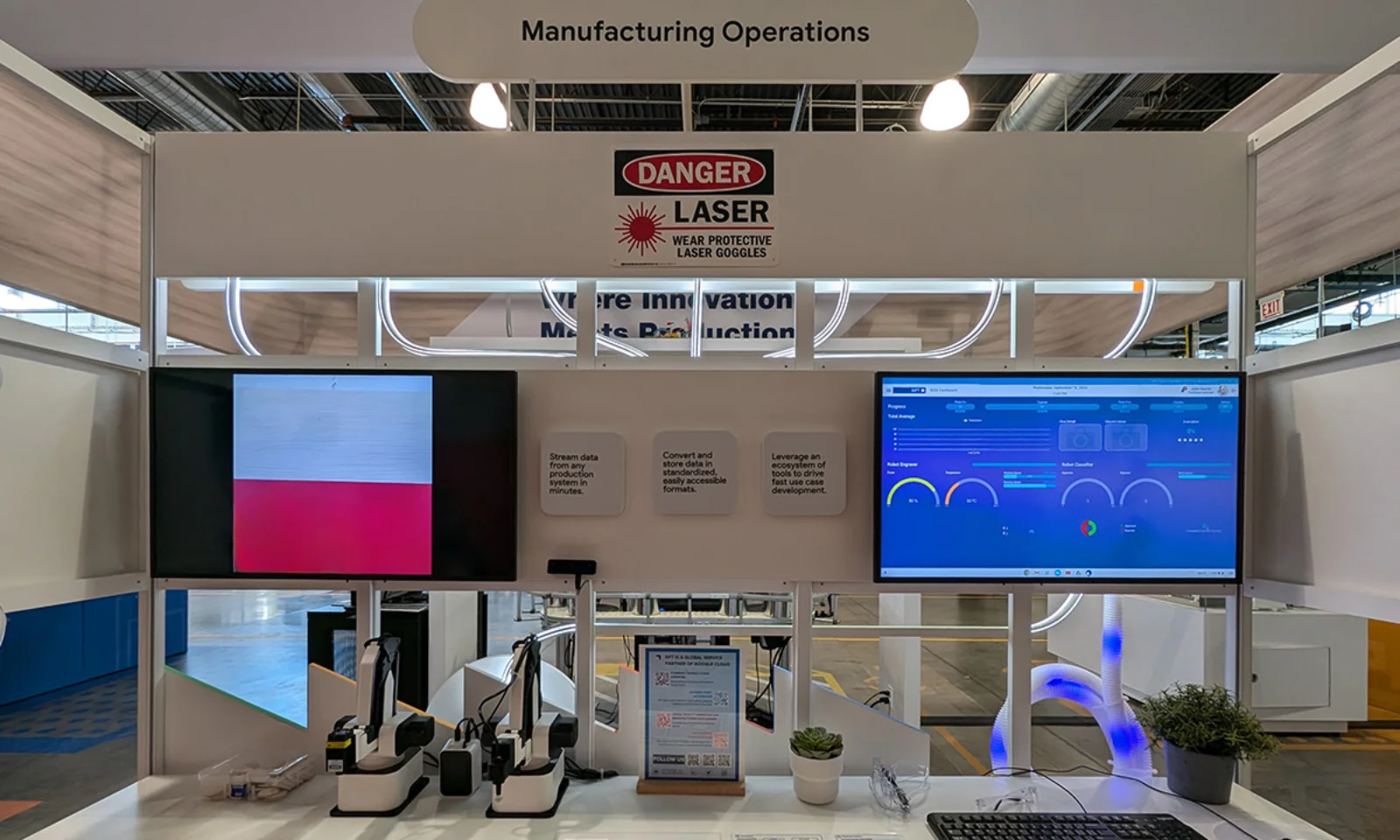  manufacturing operations display at an industrial technology exhibit, featuring two large screens showcasing data monitoring and production insights, along with laser safety warnings and connected devices on the desk.