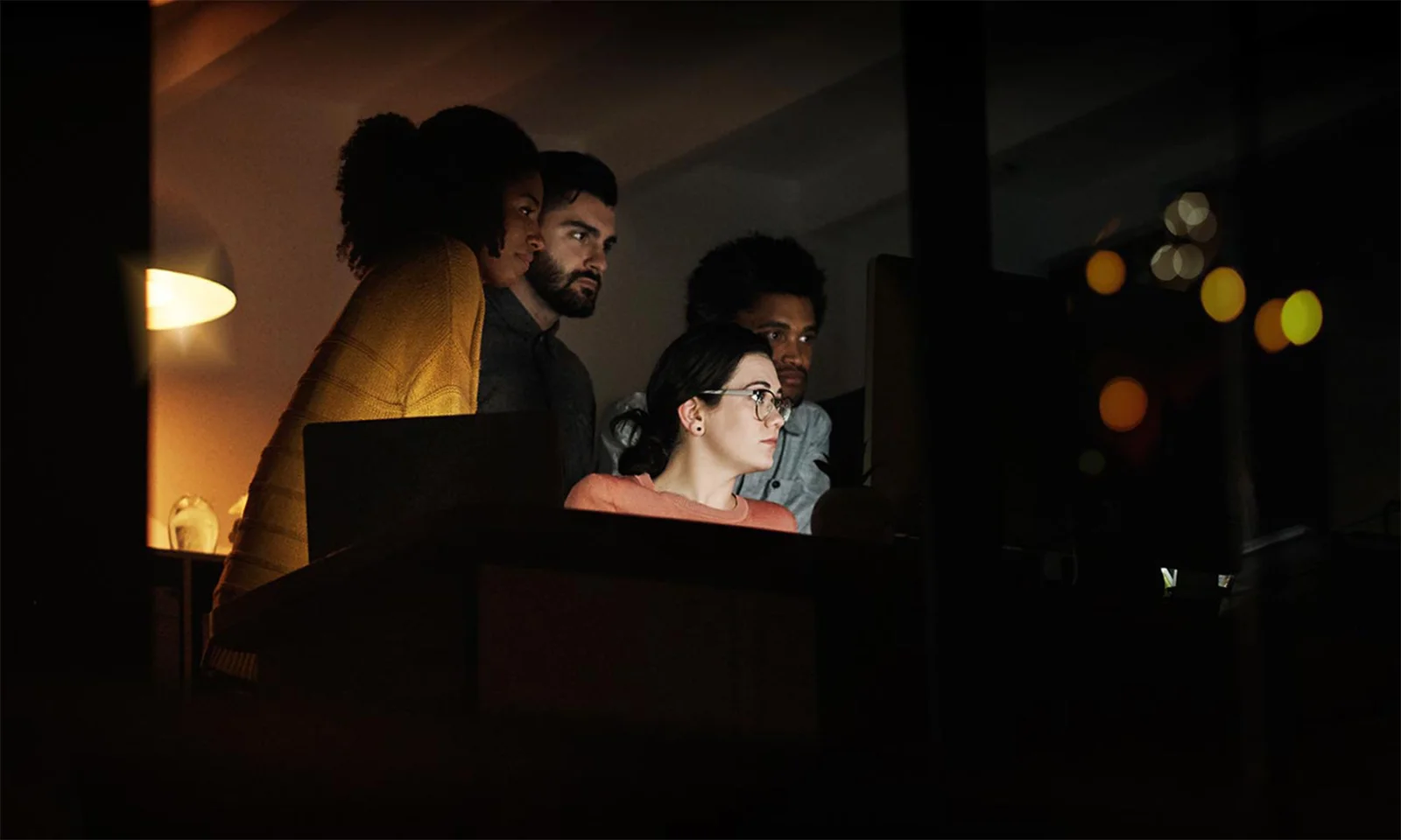 A focused team of four professionals working together on a project late at night, illuminated by the glow of computer screens in a dimly lit office.