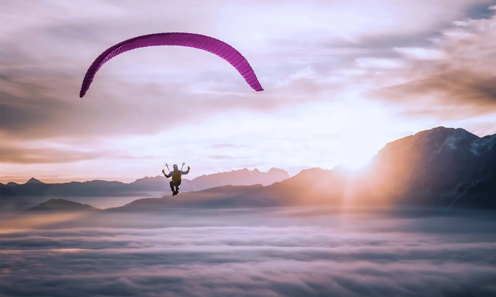 This breathtaking image captures the essence of freedom and adventure as a lone paraglider soars gracefully above a blanket of clouds. The paraglider, with a vibrant purple canopy, is set against the stunning backdrop of a sunrise, casting a warm, golden glow over the scene. The distant mountain peaks rise above the cloud layer, adding depth and a sense of scale to the image. The serene and awe-inspiring atmosphere highlights the beauty of paragliding and the unique perspective it offers on the world. This visual perfectly encapsulates the thrill and tranquillity of flying in the open sky.