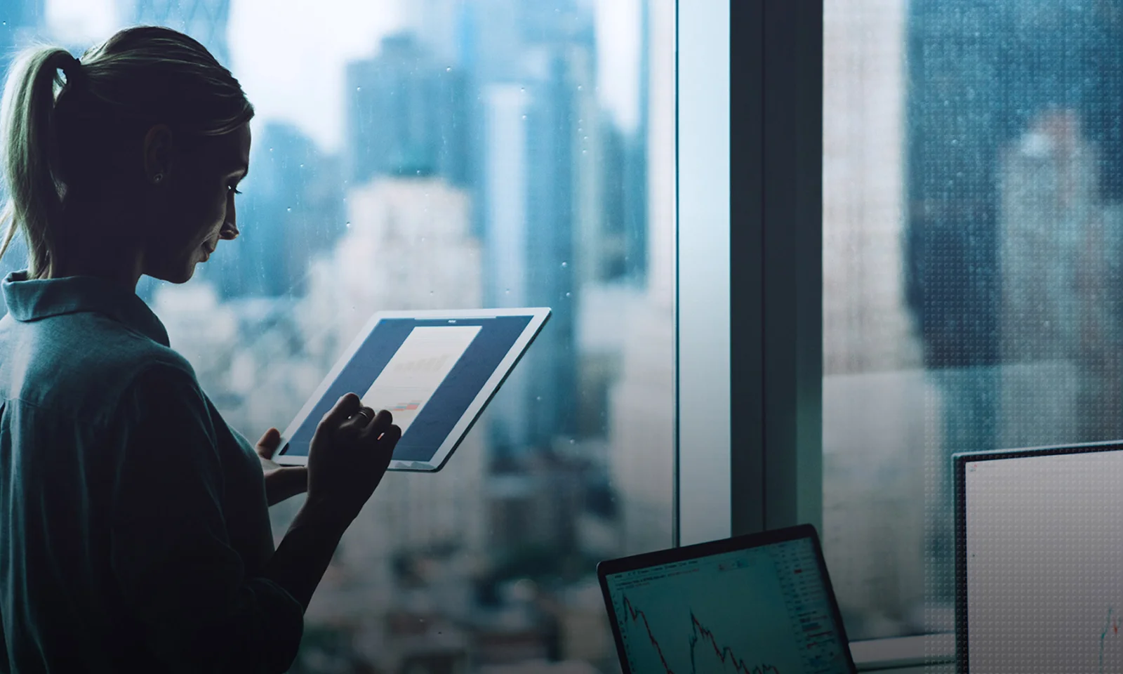 Woman analyzing financial data on a tablet while standing near a window with a cityscape background, showcasing digital transformation in business environments.