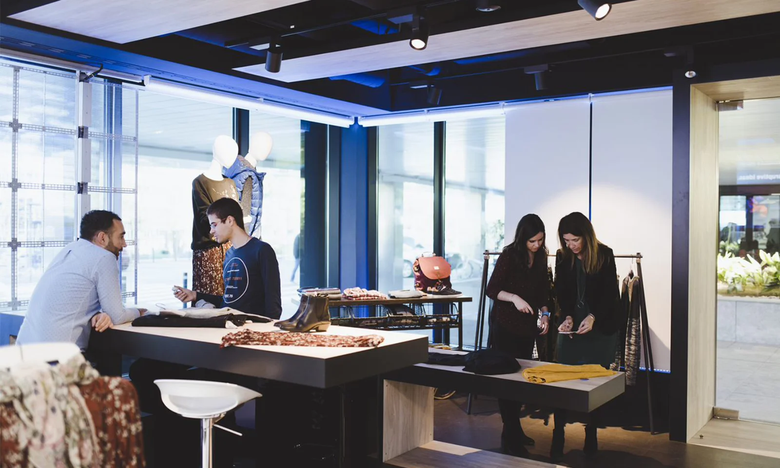 This image captures the dynamic environment of an innovation lab retail shop. Several individuals are seen engaging in collaborative activities, exploring retail technology and solutions. The setting features mannequins displaying fashionable clothing, modern retail fixtures, and various pieces of apparel laid out on tables. The bright, well-lit space with large windows fosters a creative and innovative atmosphere, ideal for brainstorming and testing new retail concepts.