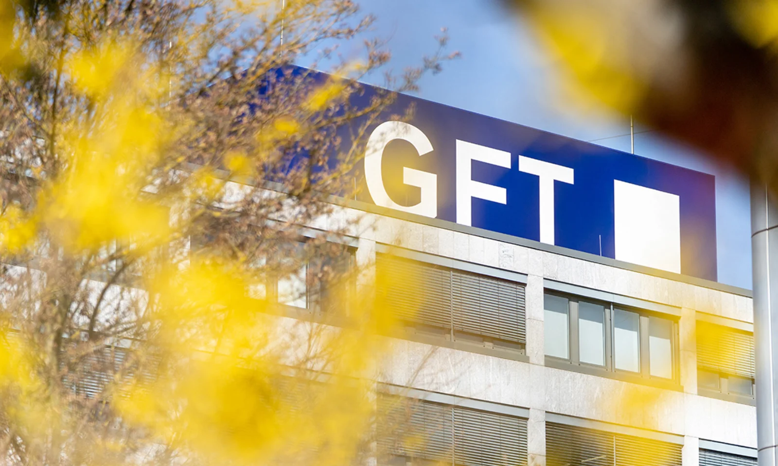 Bâtiment du siège social de GFT avec le logo de la société bien visible au sommet, encadré par un feuillage jaune vif au premier plan.