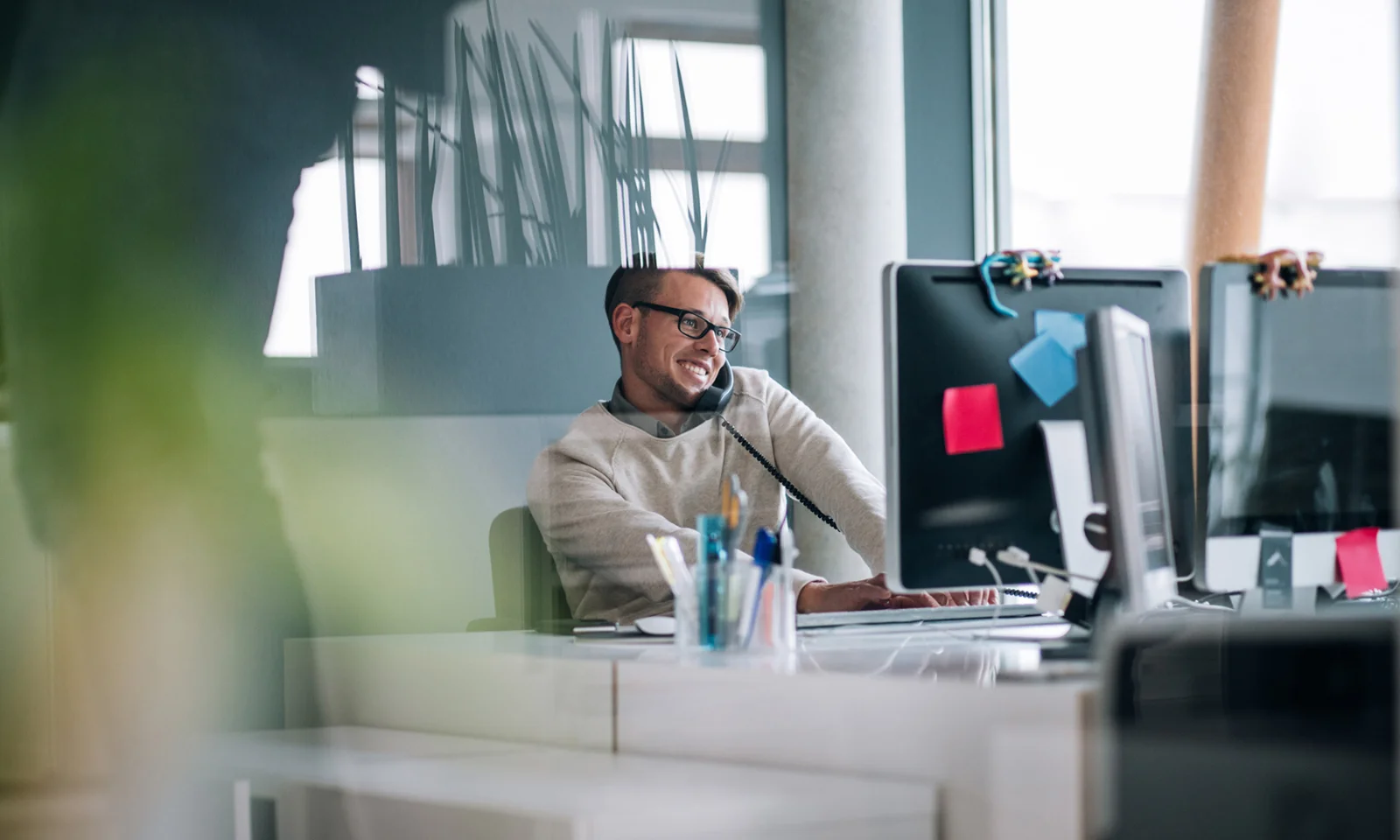 Smiling man on the phone working at a computer desk in a modern office.