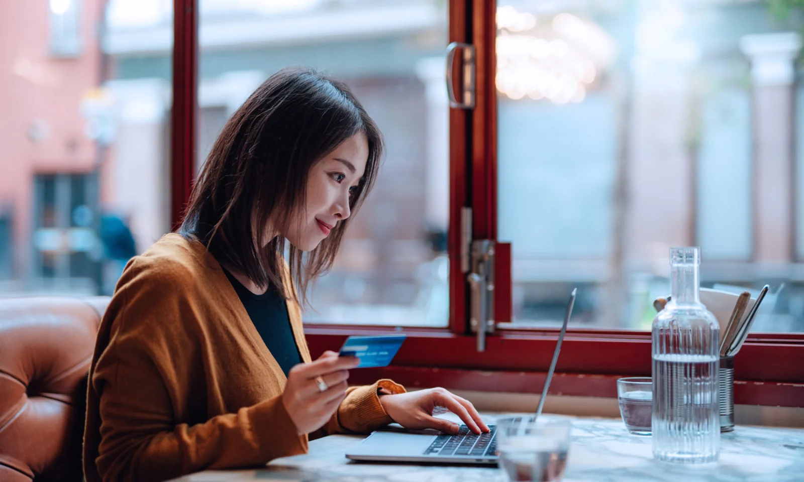 A woman holding a credit card while using a laptop in a cosy café setting, symbolising digital banking and convenience.