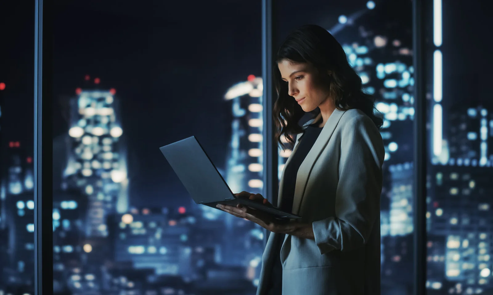 Female professional working on a laptop in a high-rise office at night, with a city skyline illuminated in the background.