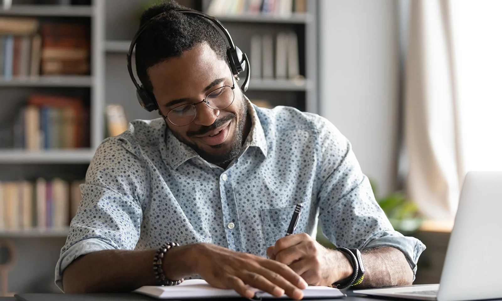 A man with headphones taking notes while listening to a podcast on his laptop, representing GFT&#039;s podcast library. He is seated in a cozy home office with bookshelves in the background, showcasing a comfortable and productive environment for learning and engagement.