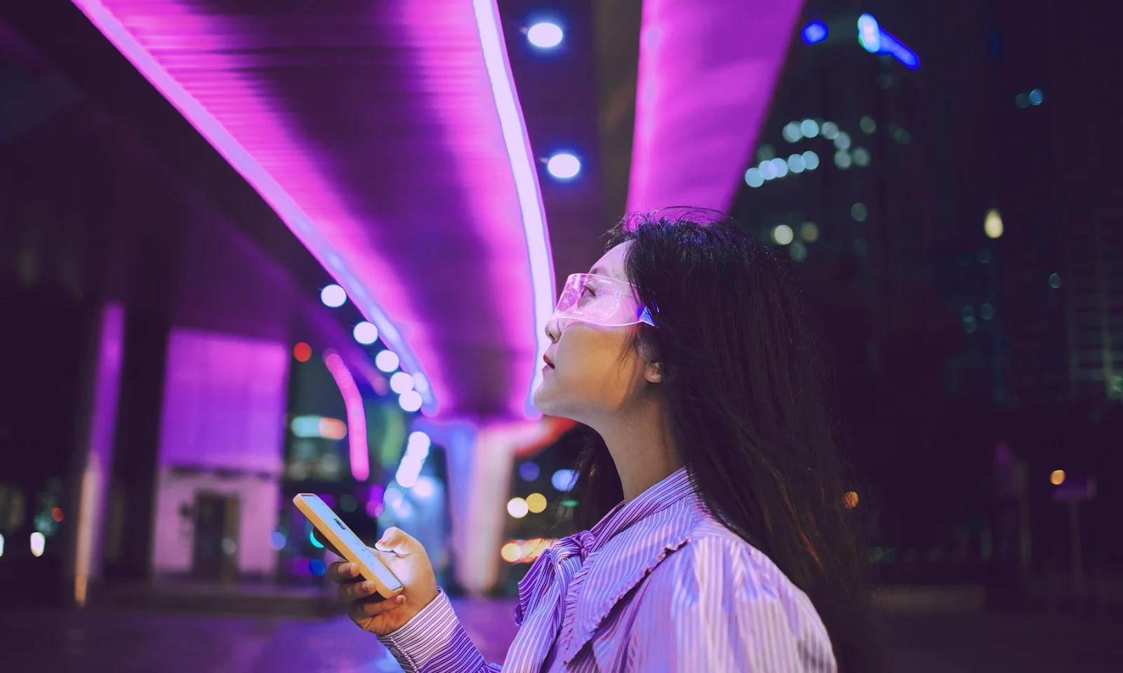 A woman wearing futuristic glasses and holding a smartphone, illuminated by purple city lights, representing the integration of AI technology into modern life.