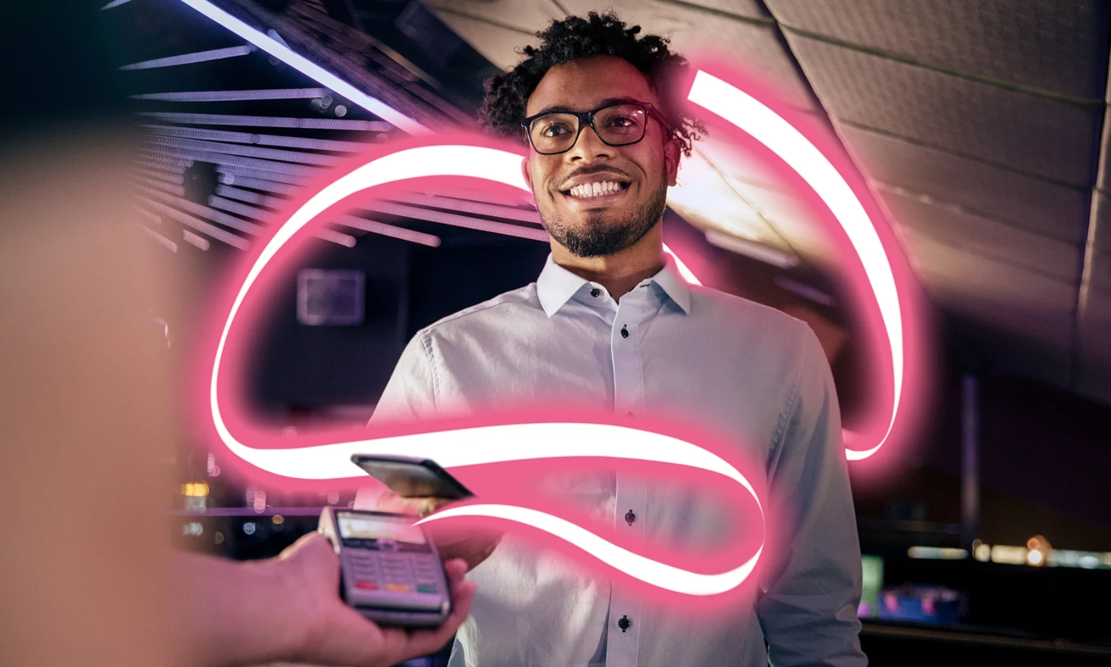 The image features a smiling man in a white shirt and glasses, standing in a modern indoor setting, making a contactless payment with a smartphone. A glowing pink light swirls around him, creating a dynamic and futuristic visual effect. The background is dark, with subtle lighting and reflections, emphasizing the man and the glowing light. This visual symbolizes modern, seamless, and innovative financial transactions, reflecting themes of advanced technology and convenience in core banking solutions.
