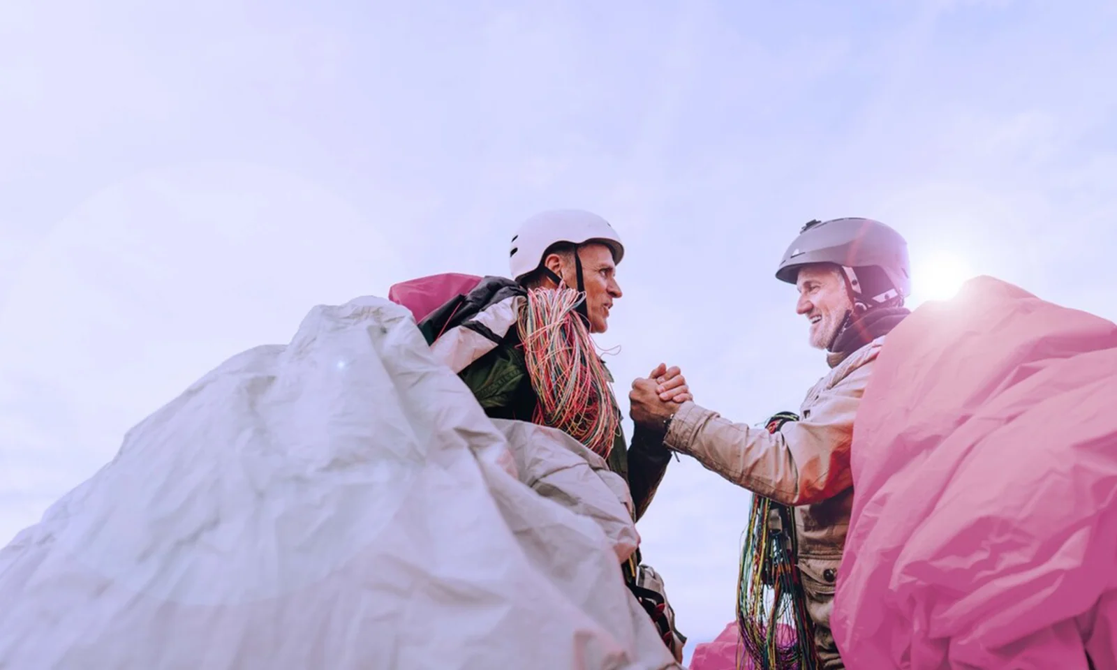 This image showcases two paragliders, moments before their takeoff, shaking hands in a gesture of camaraderie and mutual trust. Both are geared up with helmets and harnesses, their colourful parachutes draped around them, adding vibrant hues to the scene. The sky above is clear, symbolizing perfect conditions for their adventure. The image captures the essence of adventure sports—preparation, trust, and shared excitement. The mutual handshake highlights the bond and confidence between the two individuals, underscoring the human element in extreme sports.