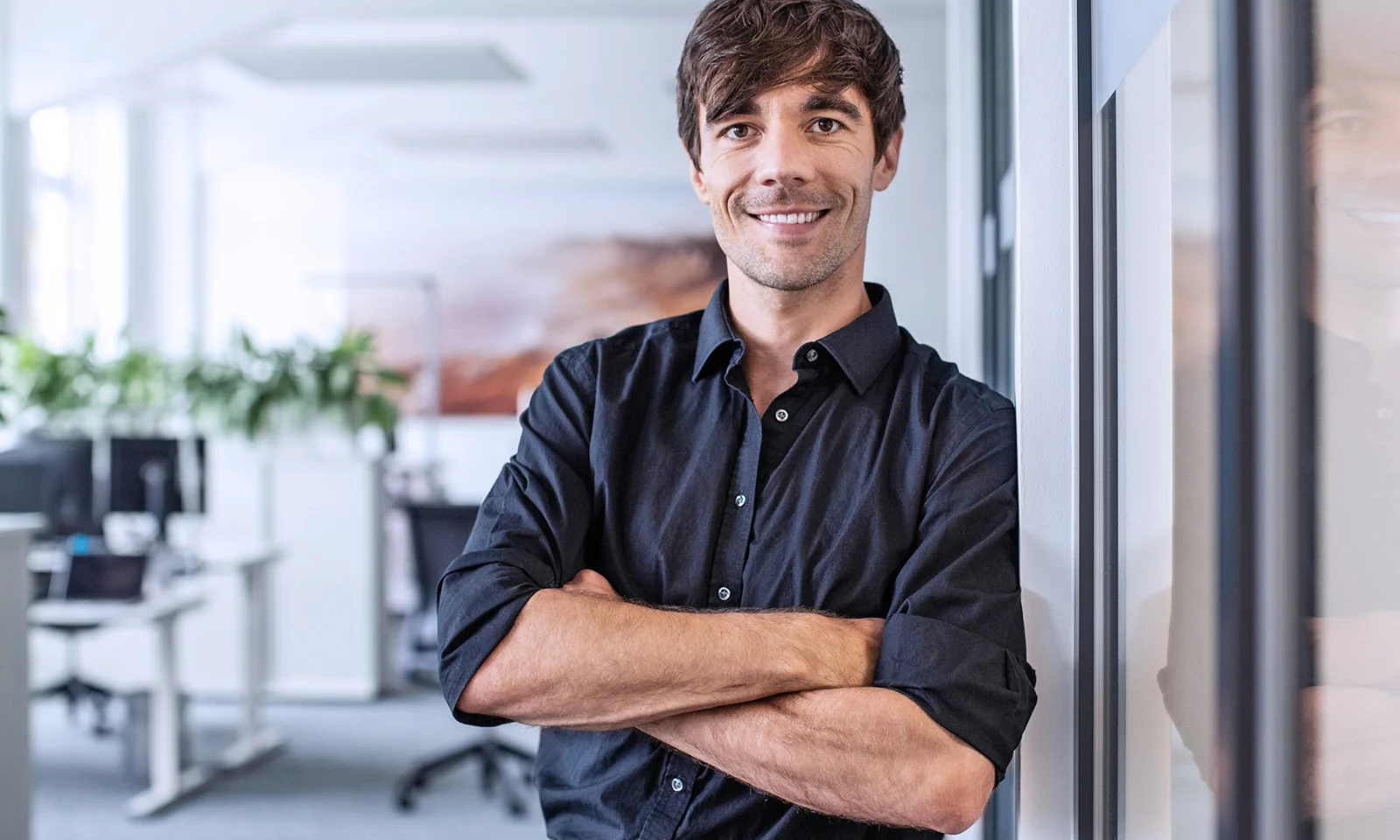 Professional man in a black shirt standing with arms crossed in an office environment.