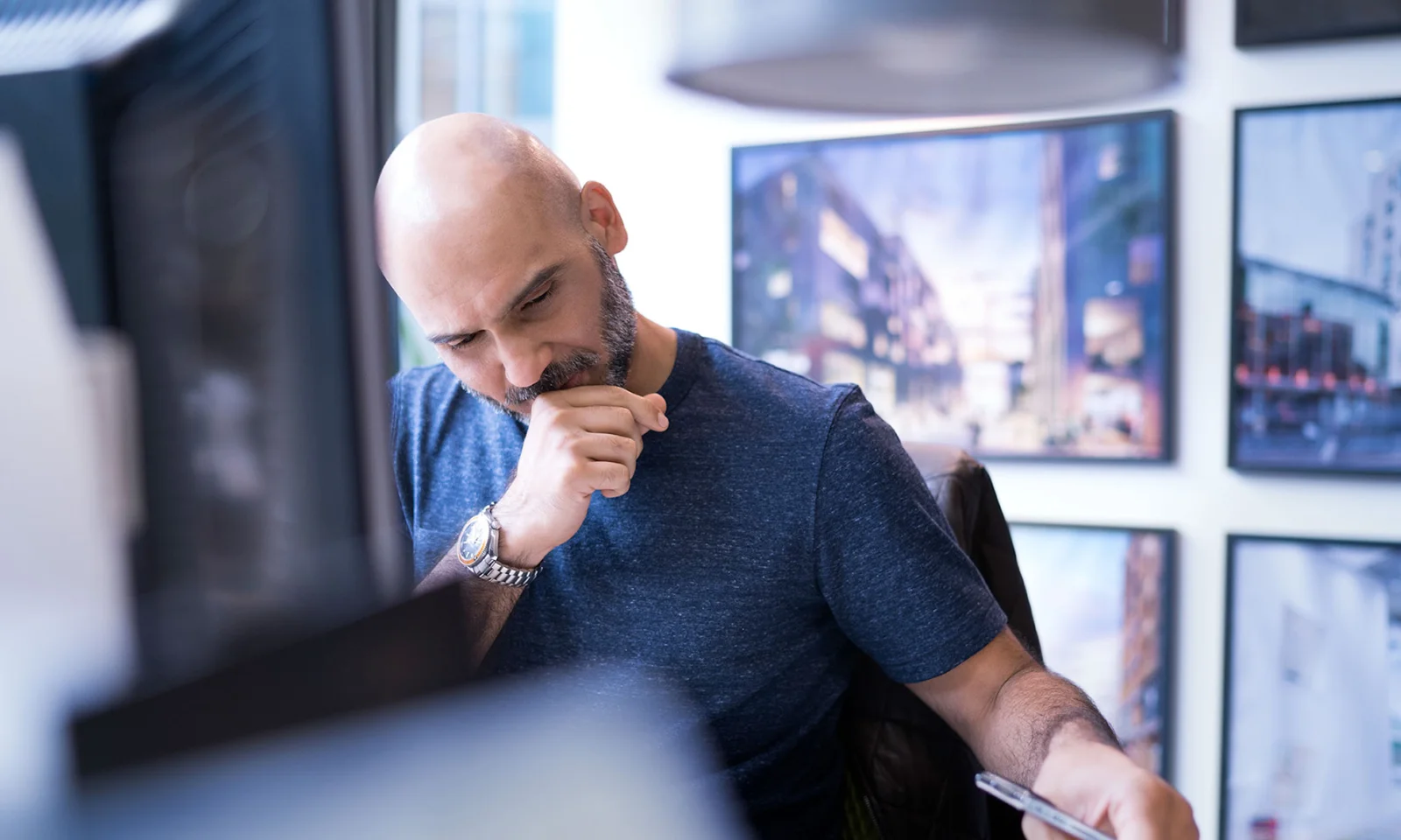 A focused professional man with a bald head and beard, wearing a blue shirt, thinking deeply while holding a pen and looking at documents on his desk.