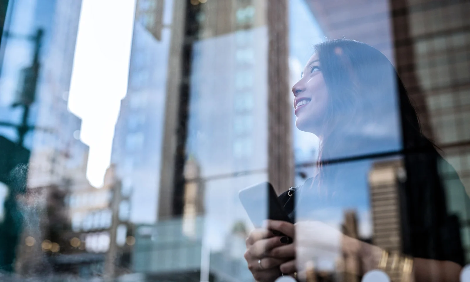 Une femme tenant un smartphone, souriant tout en levant les yeux, avec le reflet de gratte-ciels sur une vitre, symbolisant l&#039;innovation et la croissance.