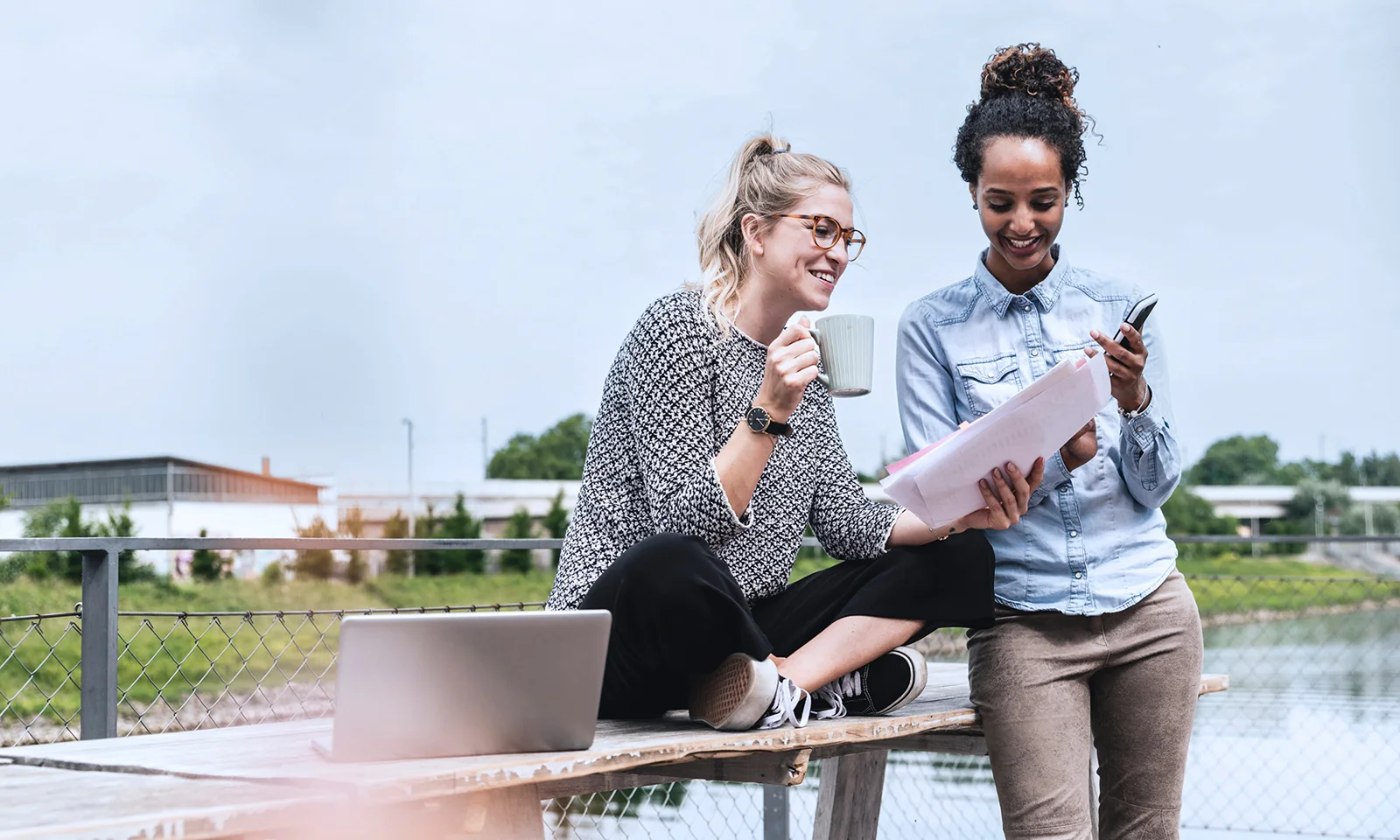 Two young women sitting outdoors, one holding a cup and the other holding papers and a phone, with a laptop on the table.