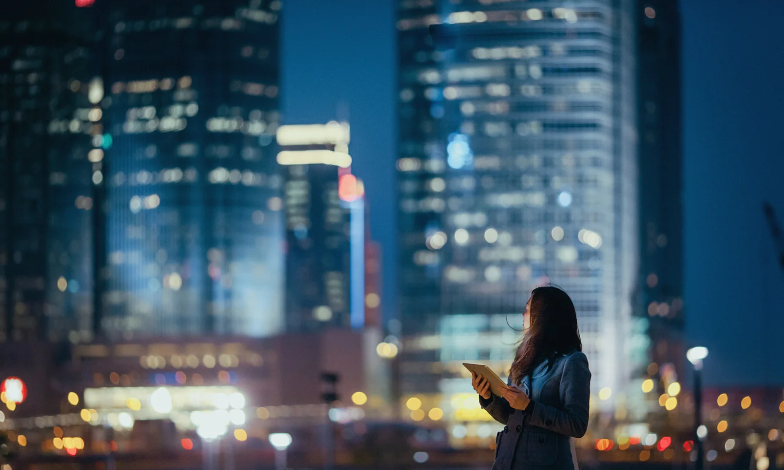 A woman holding a tablet looks up at illuminated skyscrapers in a city at night, symbolizing digital innovation and technology in an urban environment.