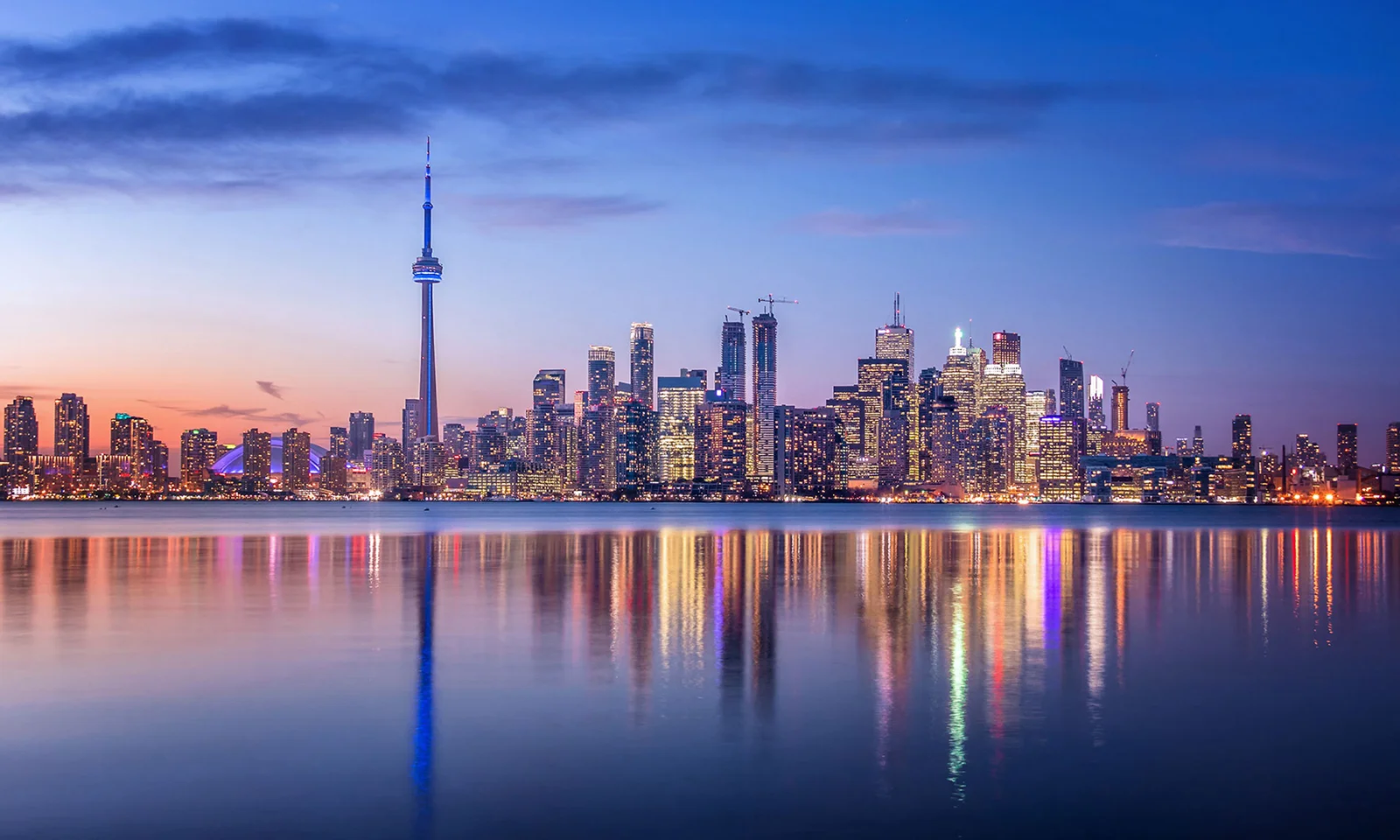The image features a stunning city skyline at dusk, with tall buildings and the iconic CN Tower prominently visible. The serene water in the foreground reflects the vibrant lights of the city, creating a symmetrical and tranquil scene. This visual captures the essence of stability, growth, and the future, making it an ideal representation for the insurance industry. The clear, calm waters and the well-lit cityscape symbolize the industry&#039;s promise of security, protection, and forward-thinking solutions.