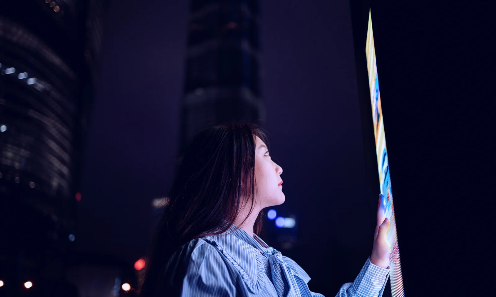 This key visual represents an overview of various industries through the imagery of a woman engaging with a large, illuminated digital screen at night. The cityscape in the background, with its towering buildings and illuminated windows, highlights the urban and industrial context. The image encapsulates the intersection of technology and modern industry, emphasizing innovation, connectivity, and the digital transformation across sectors. It serves as a visual metaphor for the broad scope and dynamic nature of various industries in the contemporary digital landscape.