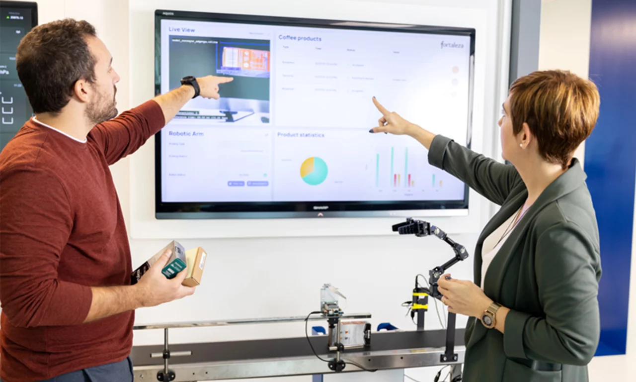 This image captures two professionals in an innovation lab industry space, engaged in a discussion. Both are pointing at data and visualizations displayed on a large screen. One holds packaging boxes, suggesting a focus on product analysis or production processes. The screen shows live views, product statistics, and other analytics related to industrial automation. The presence of a robotic arm and a conveyor belt further emphasizes the integration of advanced manufacturing technologies and data analytics in this collaborative environment. This setup reflects the modern approach to industrial innovation, leveraging digital tools for enhanced productivity and decision-making.